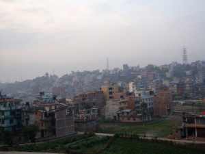 Tôt le matin, nous quittons la ville et sa vallée dans un petit car. Il faut éviter le blocus organisé par les extrémistes maoïstes (contre le gouvernement, maoïste modéré). Le paysage de pentes escarpées avec des maisons partout change radicalement dès qu'on franchit le col. Désormais, les flancs de montagnes sont taillés pour la culture en étages avec peu de végétation. Je me sens brusquement passer de la ville à la campagne, sortir de la brume et quitter la pollution. Je ressens une certaine tranquillité grâce à la nature qui surgit. Sur la route de montagne, notre petit autocar suit de haut la rivière Kitchouli (qui prend sa source au Tibet) puis une autre plus petite : Ravanste, dans la vallée d'à côté. Leur débit d'eau est faible, elles sont dans leur lit entourées de bandes de cailloux, qui seront comblées à l'époque de la mousson. La route est toujours à flanc de montagne et les terrasses autour sont rarement cultivées car l'aridité du sol ne le permet pas souvent. La production du Népal est pourtant à 90% agricole. Des petites maisons bordent sans interruption le chemin. Elles ont rarement un étage. La devanture sert souvent pour exposer des produits de commerce. On peut penser qu'une minuscule arrière-salle possède des matelas pour toute la famille. Le toit est fait de tôle ondulée parfois avec quelques pierres aux extrémités pour le maintenir. Une avancée permet d'abriter la cuisinière, une table de terre cuite en dessous de laquelle on fait un feu de bois. Plus loin, devant la maison, un tuyau et un bac permettent de se laver ou de faire la vaisselle devant les passants. Souvent, un petit abri de chaume protège une vache ou quelques chèvres. Le devant des maisons est dégagé mais quelques déchets s'amoncellent entre ou derrière, et dans la nature environnante. Des chiens sales vadrouillent dans les parages. Et partout, les femmes sont aux fourneaux avec les enfants qui aident (l'école n'étant pas obligatoire) et les hommes parfois à proximité, parfois à discuter entre eux, parfois en promenade ou éventuellement à travailler au champ ! Dans les vallées plus distantes de la route, les maisons forment des petits villages qui paraissent un peu plus riches. Les habitations sont sur deux étages. On y remarque moins le commerce et plus l'activité agricole, très visible au creux des vallées où les petites plaines sont utilisées pour la riziculture, plus commode que les terrasses sur les pentes.Des villes modestes offres des marchés avec toujours les étals en façade des maisons proposant des fruits, des légumes des viandes et des poissons exposés à l'air libre (entourés de quelques mouches qu'on chasse sans relâche). C'est rassurant de voir ces produits « frais » remplacer les canettes de soda ! Leurs habitants y sont plus souvent habillés de manière traditionnelle. Comme à Kathmandou, les rues sont en terre battue le plus souvent et parfois en macadam usé et morcelé. Les câbles électriques sont innombrables et représentent généralement le seul symbole de modernité. Ils côtoient des arbres aux fleurs mauves qui apportent une touche apaisante à la rue. Même en bordure de la route, cette flore est typiquement tropicale : les branches de arbres ont une courbure harmonieuse, enchevêtrées aux pousses de bambou géantes et variétés de citronniers géants sans fruit (pardonnez mes lacunes en botanique, je suis un citadin émerveillé). Aucun arbre ici ne ressemble à ceux que nous avons l'habitude de voir dans les zones tempérées d'Europe ! Notre bus s'est arrêté le matin à proximité d'un restaurant penché sur le vide. Devant son escalier, on remarque à peine le petit autel hindouiste avec une icône mouchetée de peinture. Nous prenons le thé sur la terrasse fleurie et baignée de soleil matinal et repartons. Le midi, il fait déjà nettement plus chaud. Nous nous arrêterons à un restaurant ouvert de toutes parts, au milieu d'un jardin à la japonaise où les fleurs arborent de multiples couleurs. Y faire une sieste aurait été un bonheur, mais nous craignons l'extension du blocus des maoïstes à plusieurs routes et devons reprendre le chemin pour Pokhara, la ville d'où nous devons commencer notre marche dans les montages. Des policiers tiennent à nous « escorter » (ouvrir la route et nous accompagner), mais leur aide n'est pas très efficace, nous nous arrêtons dans une petite ville, la route est bloquée plus loin. A s'y promener, on remarque que la rue principale est très animée concentrant les marchés, mais plus on s'en éloigne, plus les rues sont désertes. Le car repart. En sortant de la ville, on observe un rassemblement de maoïstes (gouvernementaux ceux-là) organisé sur le terrain -vague- de sport, beaucoup y affluent machinalement. Les abords de la ville de Pokhara (au Nord-Ouest de Kathmandou, au pied des Annapurnas) sont relativement riches par rapport à ce que nous avons vu jusqu'à présent. Les habitations sont plus grandes, ont plusieurs étages et sont plus espacées dans les rues. Nous nous arrêterons juste sur un parking pour accueillir nos cinq porteurs, qui accompagneront notre randonnées ces dix prochains jours, et porteront le gros de nos bagages ! Je suis le seul à le découvrir et m'en étonner. Mais il faut se plier aux règles du groupe. Ayant rassemblé leminimum d'affaires dans un sac de randonnée, je suis pourtant mal à l'aise d'avoir des « domestiques ». Néanmoins, ces jeunes accompagnateurs locaux sauront probablement m'éclairer sur les coutumes du pays d'une manière différente de celle de notre guide. Une fine pluie commence à tomber et le départ de notre « trek » n'est plus très loin ! Je suis plus qu'impatient de pouvoir enfin quitter la route, entrer dans la nature profonde, fouler les flancs de montagnes et véritablement commencer l'aventure. Lorsque notre car arrive, la pluie s'arrête subitement. De notre groupe, chacun se prépare alors avec minutie: c'est impressionnant de voir l'équipement des randonneurs d'aujourd'hui : chaussures-chaussons, bâtons de ski, t-shirt anti-transpiration, sacs à dos ergonomiques avec une gourde munie d'un tuyau à portée de la bouche pour se déshaltérer sans s'arrêter… je dois faire pâle figure avec mon matériel de récupération, mais notre jovial doyen, Jean-Claude, esquisse un sourire, il se contente lui aussi de matériel rustique. Les porteurs, eux, ont de simples baskets ! Même avec leur technologie de pointe, certains s'inquiètent de devoir monter pendant deux heures sans échauffement ! C'est là que j'ai compris que tous ne sont pas venus ici pour une quête intérieure. Le parcours se fait sur des escaliers d'ardoises entre les champs en étages, à traverser quelques minuscules villages qui proposent – eux aussi – des sodas et des paquets de chips. Il y a aussi les premiers drapeaux à prières et quelques arbres rhododendrons, très exotiques. Mon estomac commence à me jouer des tours, sans doute dû à la nourriture, ou à mon empressement de gravir l'Himalaya. Qu'à cela ne tienne, le paysage est si impressionnant qu'on en oublie presque tout le reste. Notre groupe s'arrête au soir à Dhampus. La lodge est formée de deux gros bâtiments à étages. En plus des habitants qui s'occupent de la cuisine et de l'intendance, il y a là un groupe de japonais suréquipés… Nous ne sommes pas les seuls visiteurs étrangers ici. Après le thé, s'ensuit le ballet des douches (tièdes voire froides mais il fallait être dans les premiers). Au repas, les assistants de notre guide nous servent des « momos », des raviolis fourrés aux légumes, cuits à la vapeur. Il y a un feu de camp dehors, certains de notre groupe se joignent au petit cercle. On ne distingue pas bien qui est présent, ce sont probablement nos porteurs avec qui nous échangeons quelques timides regards dans la pénombre. Peu de discussion, la chaleur du feu et le rythme de quelques tambours suffisent à nous faire apprécier cette première nuit ensemble dans les montagnes.